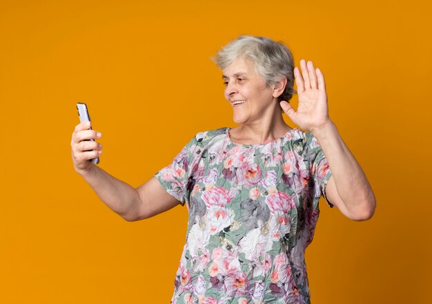 Sonriente anciana levanta la mano sosteniendo y mirando el teléfono aislado en la pared naranja