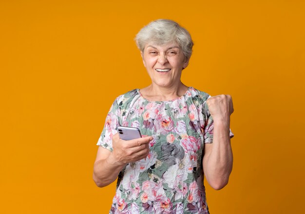 Sonriente anciana mantiene el puño sosteniendo el teléfono aislado en la pared naranja