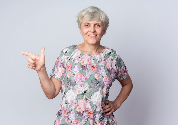 Sonriente anciana pone la mano en la cintura apuntando al lado aislado en la pared blanca