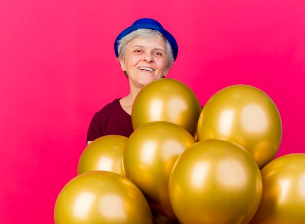 Sonriente anciana con sombrero de fiesta se encuentra con globos de helio mirando a la cámara en rosa