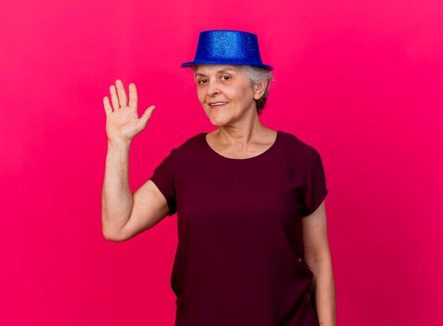 Foto gratuita sonriente anciana vistiendo gorro de fiesta se encuentra con la mano levantada en rosa