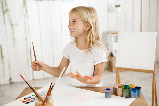 Sonriente ángel hermoso niño con cabello rubio con camiseta blanca pintando en su palma.