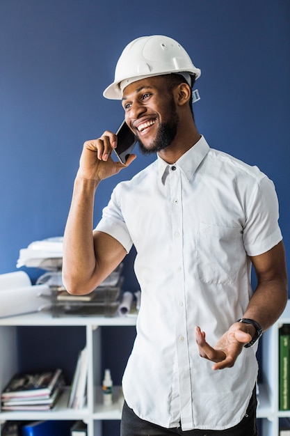 Sonriente arquitecto africano hablando por teléfono móvil en la oficina