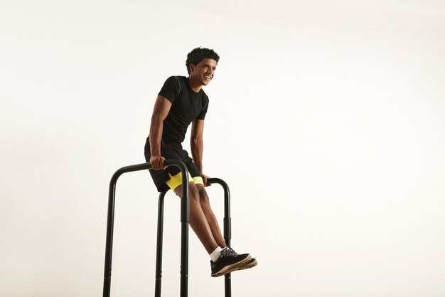 Sonriente atleta afroamericano fuerte con un afro con camisa sintética negra y pantalones cortos negros y amarillos haciendo L-se sienta en barras cortas en casa aislado en blanco.