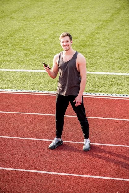 Foto gratuita sonriente atleta masculino de pie en la pista de carreras con teléfono móvil en la mano