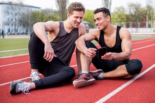 Sonriente atleta masculino sentado en la pista de carreras mostrando teléfono móvil a su amigo