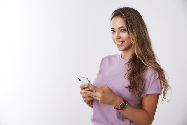 Sonriente y atractiva mujer caucásica bronceada de pelo rizado de 25 años que sostiene un teléfono inteligente a medias girando la cámara sonriendo misteriosamente quiere pedir ayuda usando una nueva aplicación de gadget de fondo blanco
