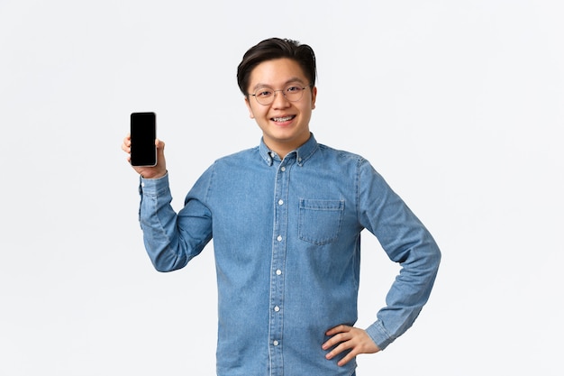 Sonriente autónomo masculino asiático satisfecho, empresario con su propia pequeña empresa que muestra la pantalla del teléfono inteligente complacido. Chico con aparatos ortopédicos y gafas mediante aplicación móvil, fondo blanco.