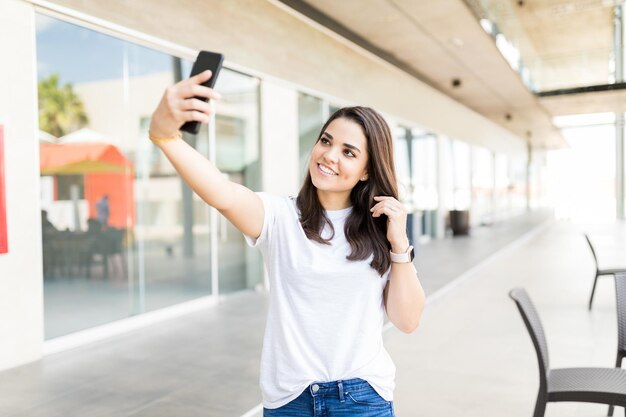 Sonriente bloguera adulta mediana tomando selfie usando un teléfono inteligente en un centro comercial