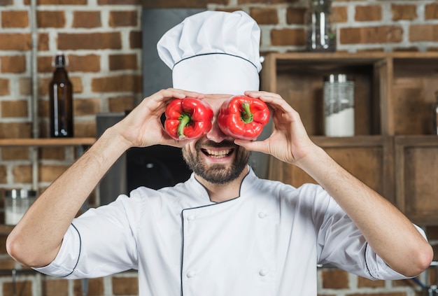 Sonriente chef hombre sosteniendo pimiento rojo delante de sus ojos