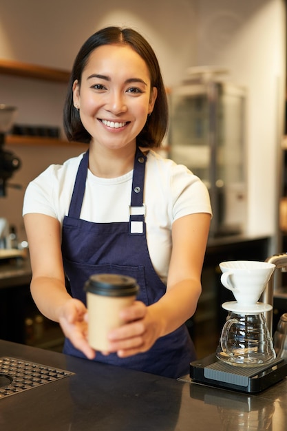 Foto gratuita sonriente chica asiática barista dando orden al cliente sosteniendo una taza de café para llevar con delantal trabajando