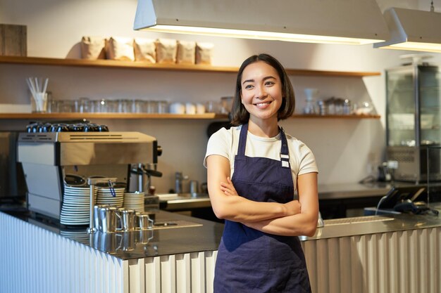 Sonriente chica asiática barista de pie cerca del mostrador en el café con delantal de la empresa cruza las manos y mira