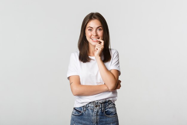 Sonriente chica atractiva mordiendo el dedo y mirando con tentación, blanco.