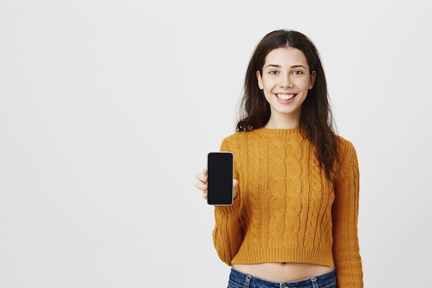 Sonriente chica atractiva que muestra la aplicación, apuntando con el dedo a la pantalla del teléfono móvil