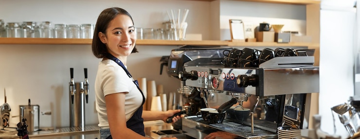 Foto gratuito sonriente chica barista asiática hace capuchino con máquina de café detrás del mostrador en la cafetería