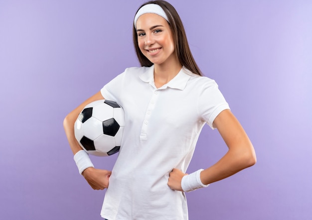 Sonriente chica bastante deportiva con diadema y muñequera con balón de fútbol poniendo las manos en la cintura aislado en el espacio púrpura