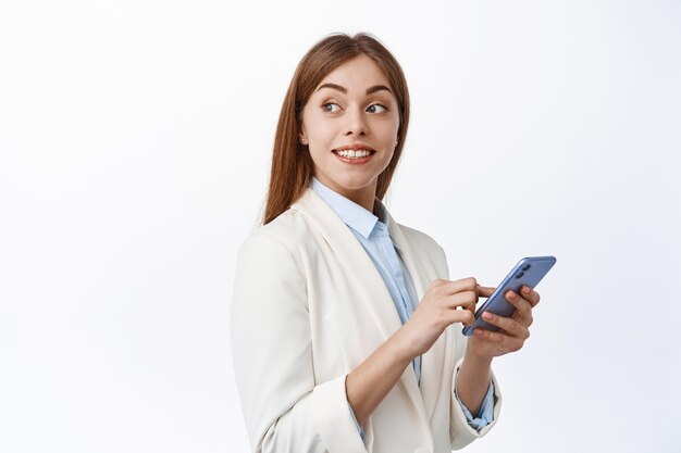 Sonriente chica corporativa en traje usando un teléfono móvil, gire la cabeza hacia atrás y mire el texto promocional, leyendo el espacio de la copia en la pared blanca, sosteniendo el teléfono inteligente