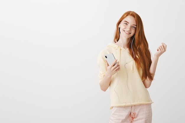Sonriente chica pelirroja escuchando música en auriculares y sosteniendo el teléfono inteligente