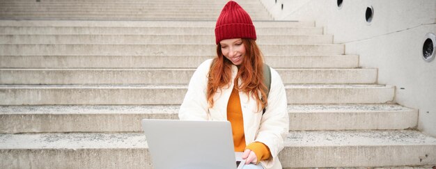 Sonriente chica pelirroja joven escribiendo en el teclado de la computadora portátil sentado al aire libre en las escaleras con la computadora