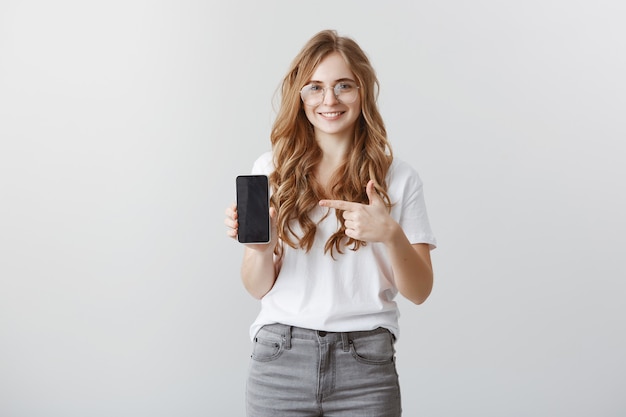 Sonriente chica rubia atractiva con gafas apuntando con el dedo a la pantalla del teléfono inteligente, mostrando la aplicación