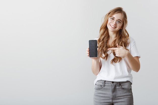 Sonriente chica rubia atractiva con gafas apuntando con el dedo a la pantalla del teléfono móvil, mostrando la aplicación