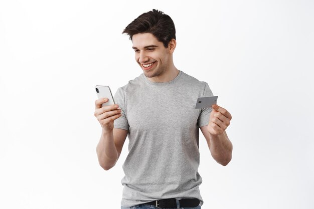 Sonriente chico casual tienda online, pagando con tarjeta de crédito y smartphone, mirando el teléfono con cara de satisfacción, compra, de pie contra el fondo blanco.