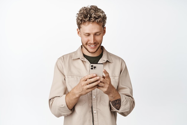 Sonriente chico caucásico con smartphone Guapo chateando en el teléfono móvil de pie sobre fondo blanco.