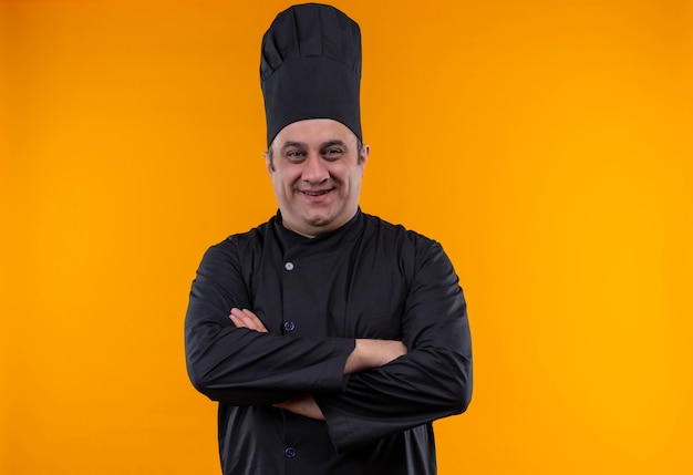 Sonriente cocinero de sexo masculino de mediana edad en uniforme de chef cruzando las manos sobre fondo amarillo