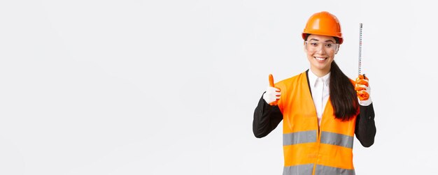 Sonriente y confiada arquitecta ingeniera de construcción asiática con uniforme de casco de seguridad que muestra cinta métrica y pulgar hacia arriba satisfecha con los buenos resultados manteniéndose bien hecho con el fondo blanco del equipo