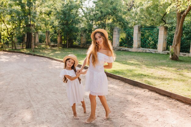 Sonriente dama delgada en vestido blanco de moda posando junto a la pequeña hija en la calle con valla de hierro. Retrato al aire libre de linda chica y su mamá delgada con sombrero, pasar tiempo en el parque.