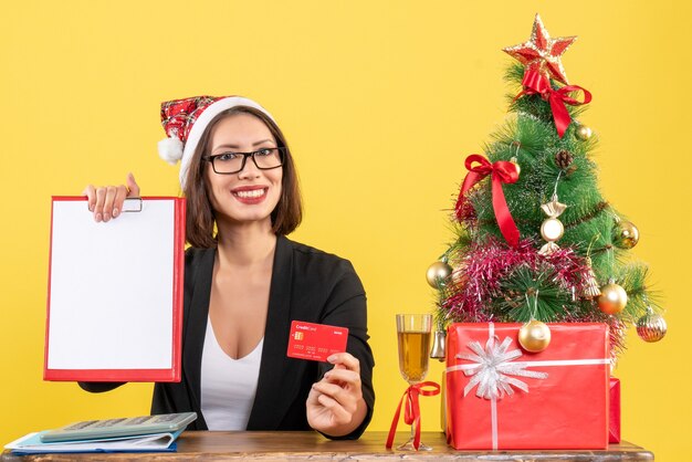 Sonriente dama encantadora en traje con sombrero de santa claus y anteojos mostrando tarjeta bancaria y documento en la oficina en amarillo aislado