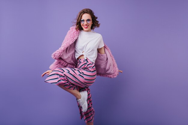 Sonriente dama europea en gafas de sol de moda bailando en chaqueta de piel. hermosa modelo de mujer con cabello castaño ondulado saltando durante la sesión de fotos.