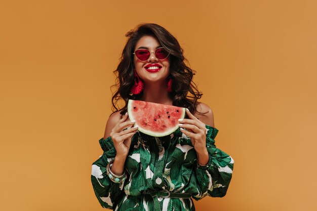 Sonriente dama de moda fresca con peinado moderno rizado en aretes rojos y ropa elegante verde posando con sandía sobre fondo naranja