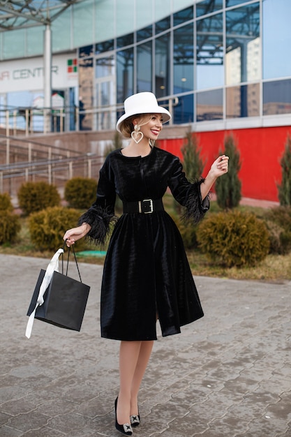 Sonriente dama muy elegante con sombrero blanco y vestido negro caminando por la calle. Concepto de calle de moda