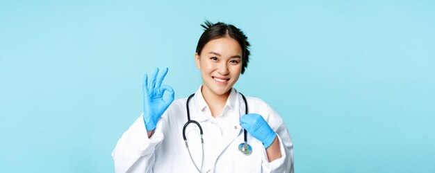 Sonriente doctora asiática doctora tocando estetoscopio mostrando bien ok firmar en aprobación fondo azul