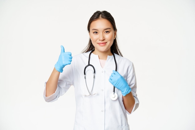 Sonriente doctora asiática muestra los pulgares hacia arriba, usa guantes de goma y uniforme de clínica, se encuentra sobre fondo blanco.