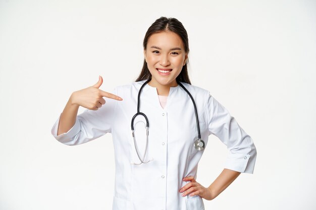Sonriente doctora asiática, trabajadora médica profesional real, señalando con el dedo a sí misma, vestida con una bata médica y un estetoscopio, fondo blanco.