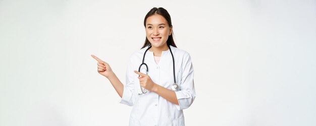 Sonriente doctora asiática en uniforme médico señalando con el dedo y mirando a la izquierda en la promoción del espacio de copia de publicidad de pie en bata contra fondo blanco