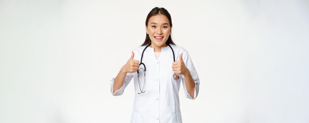 Sonriente doctora asiática vistiendo túnica médica mostrando Thumbs up complacido recomendando productos de salud