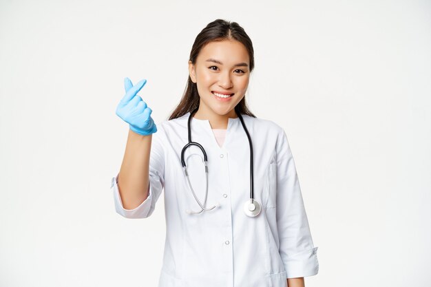 Sonriente doctora coreana muestra gesto de corazón con los dedos en guantes de goma, vistiendo uniforme médico, mirando feliz a la cámara, fondo blanco.