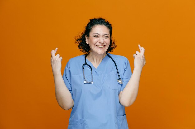 Sonriente doctora de mediana edad con uniforme y estetoscopio alrededor del cuello mirando a la cámara pidiendo deseos mientras cruza los dedos aislados en un fondo naranja