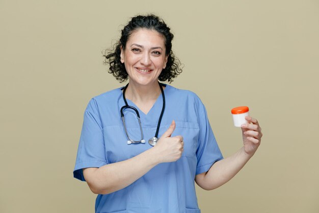 Sonriente doctora de mediana edad con uniforme y estetoscopio alrededor del cuello mostrando un contenedor de tabletas mirando a la cámara mostrando el pulgar hacia arriba aislado en el fondo verde oliva