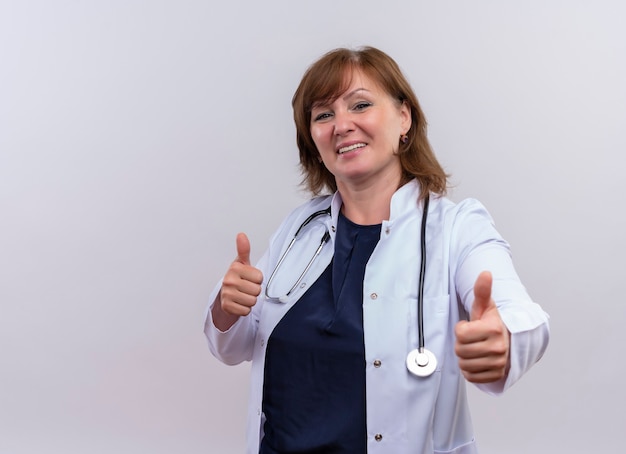 Sonriente doctora de mediana edad vistiendo bata médica y estetoscopio mostrando los pulgares hacia arriba en la pared blanca aislada con espacio de copia