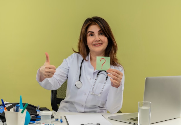 Sonriente doctora de mediana edad vistiendo bata médica y estetoscopio sentado en el escritorio con herramientas médicas y portátil con signo de interrogación mostrando el pulgar hacia arriba aislado