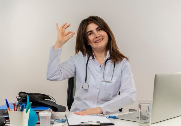 Sonriente doctora de mediana edad vistiendo bata médica y estetoscopio sentado en el escritorio con portapapeles de herramientas médicas y portátil haciendo signo ok aislado