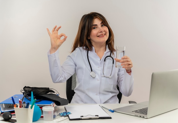 Sonriente doctora de mediana edad vistiendo bata médica y estetoscopio sentado en el escritorio con portapapeles de herramientas médicas y portátil haciendo signo de ok sosteniendo un vaso de agua aislado