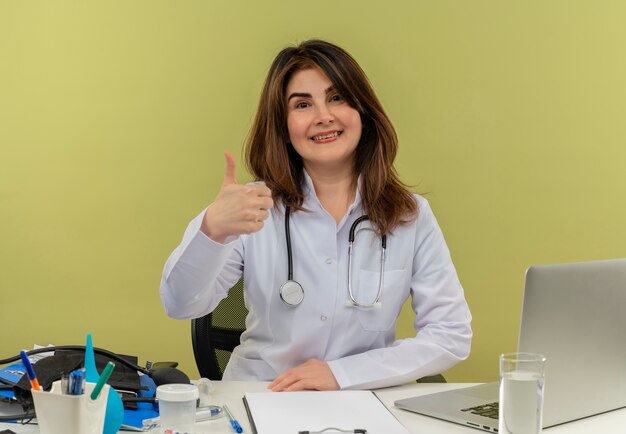 Sonriente doctora de mediana edad vistiendo bata médica y estetoscopio sentado en el escritorio con portapapeles de herramientas médicas y portátil mostrando el pulgar hacia arriba aislado