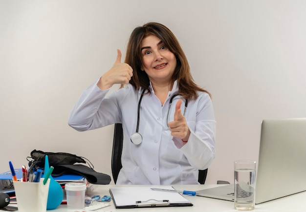 Sonriente doctora de mediana edad vistiendo bata médica con estetoscopio sentado en el escritorio trabajar en una computadora portátil con herramientas médicas que le muestran un gesto con el pulgar hacia arriba en la pared blanca