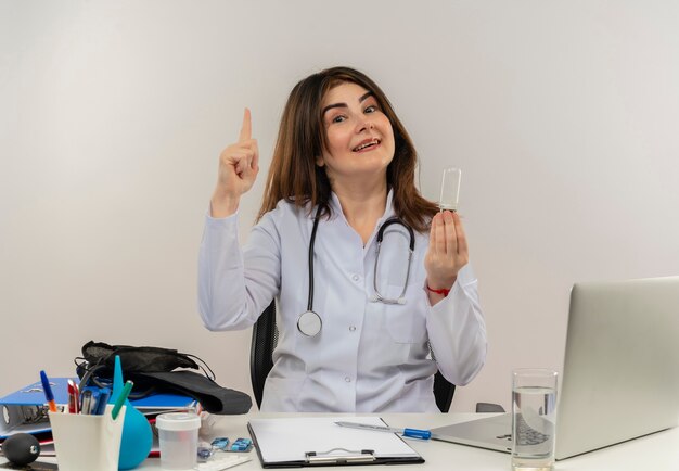 Sonriente doctora de mediana edad vistiendo bata médica con estetoscopio sentado en el escritorio trabajar en una computadora portátil con herramientas médicas sosteniendo la bombilla y apuntando hacia arriba en la pared blanca