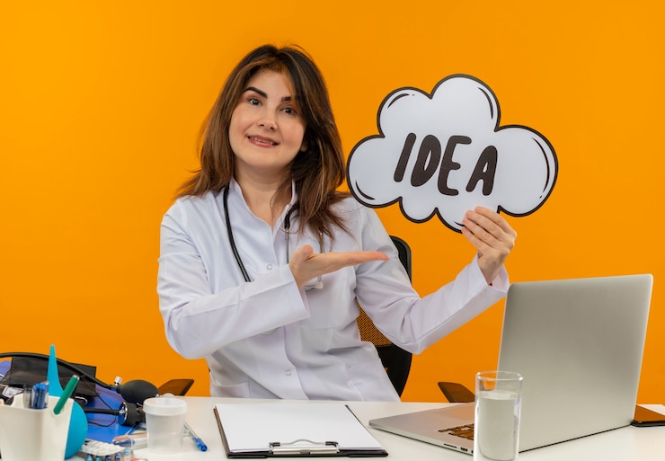 Foto gratuito sonriente doctora de mediana edad vistiendo bata médica con estetoscopio sentado en el escritorio, trabajo en computadora portátil con herramientas médicas sosteniendo y puntos con burbuja de idea de mano en pared naranja